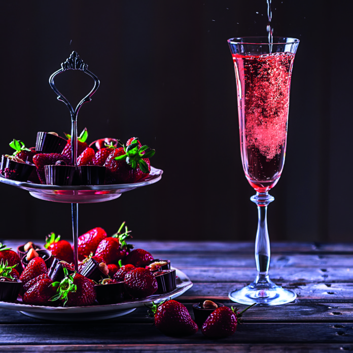Sparkling pink wine is poured in glass. Stand with strawberries and sweets on a wooden table.