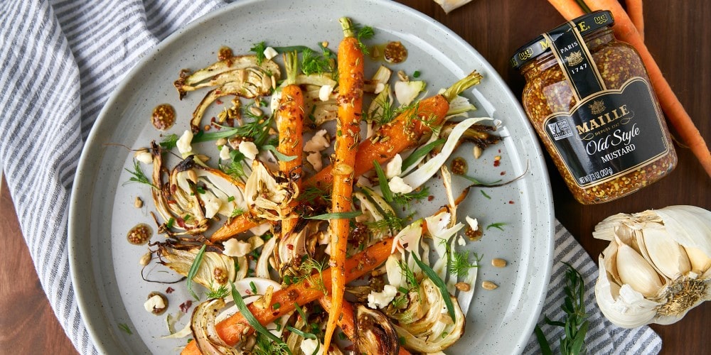 An image of roasted carrots and fennel salad on a gray plate on a striped towel with Maille Old Style