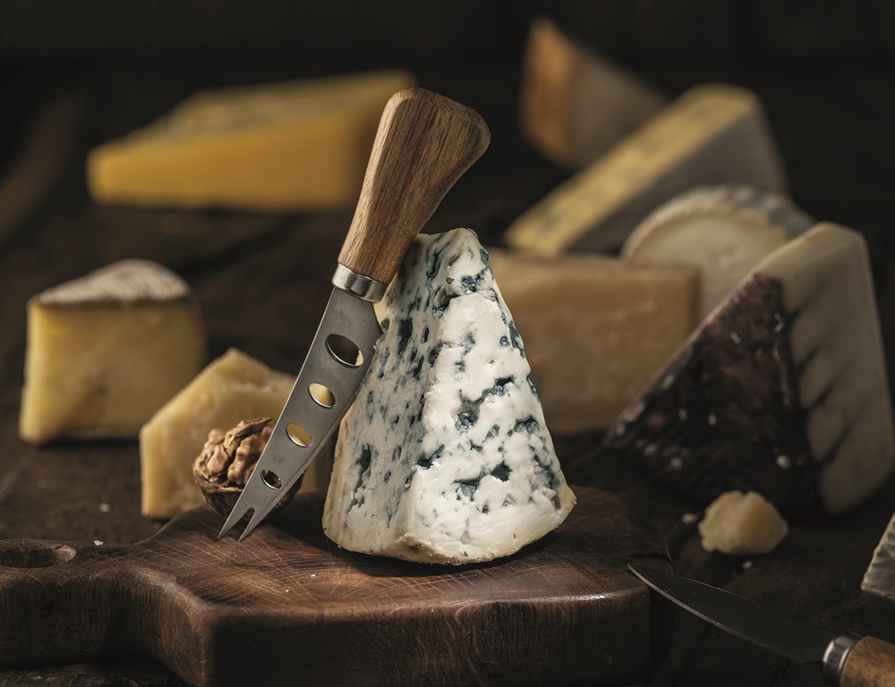 Segment of blue mould cheese and cheese knife on wooden board. Different cheeses at the background.
